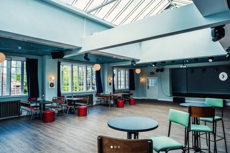 Light-filled community hall and cafe venue hire in Lewisham SE6 via Love Locations, displaying retro dining chairs, round tables, and large skylight windows.