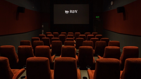Symmetrical view looking down the center aisle towards the illuminated projection screen displaying the Ritzy logo inside The Ritzy Screen 4 film location in Brixton SW2 from Love Locations, featuring plush red velvet seats.