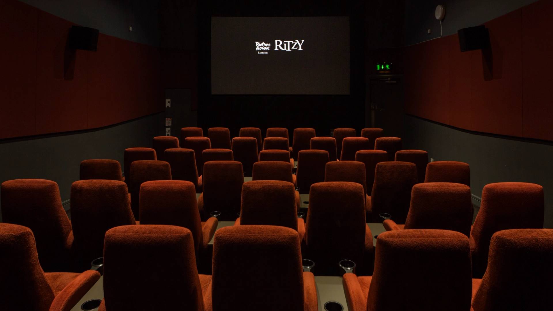 Symmetrical view looking down the center aisle towards the illuminated projection screen displaying the Ritzy logo inside The Ritzy Screen 4 film location in Brixton SW2 from Love Locations, featuring plush red velvet seats.