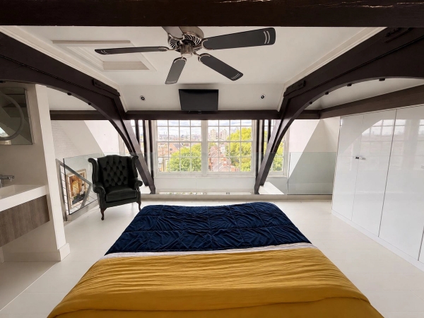 Characterful loft bedroom location house in Brixton SW4 by Love Locations, featuring sloping rooflines, dark structural beams, a ceiling fan, and large school sash windows.