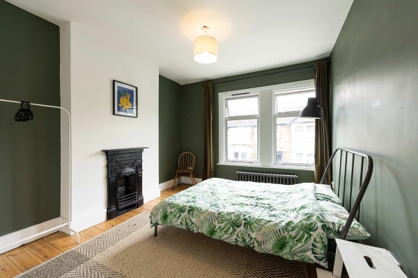 Love Locations characterful bedroom space East London: Sunlit bedroom featuring an original black cast iron fireplace hearth, rustic wooden floors, and expansive windows, ideal for editorial photography.