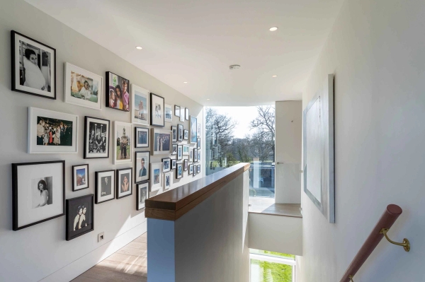 Love Locations architectural staircase location Godalming Surrey GU8: Light-flooded upper landing area featuring structural glass balustrades and an extensive grid gallery wall of framed prints.