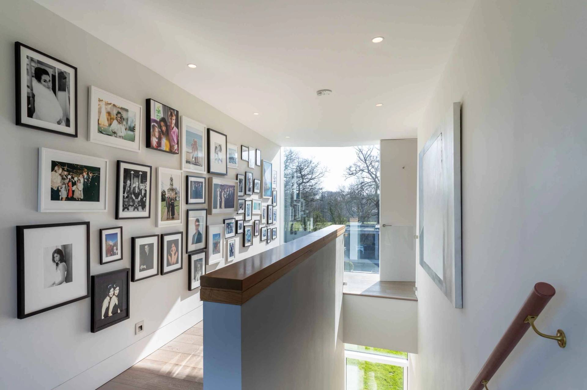 Love Locations architectural staircase location Godalming Surrey GU8: Light-flooded upper landing area featuring structural glass balustrades and an extensive grid gallery wall of framed prints.
