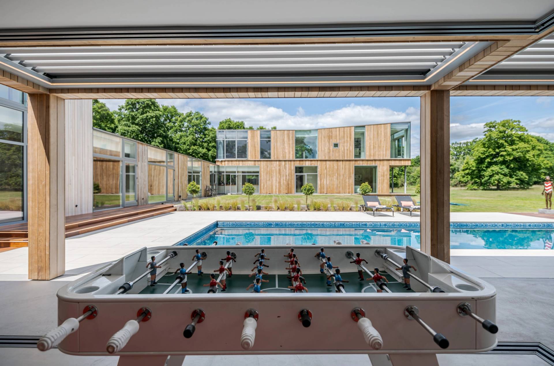 Love Locations luxury outdoor pool location Surrey: Looking past a courtyard foosball table toward a pristine open-air swimming pool and contemporary timber architecture, excellent for summer commercial ads.