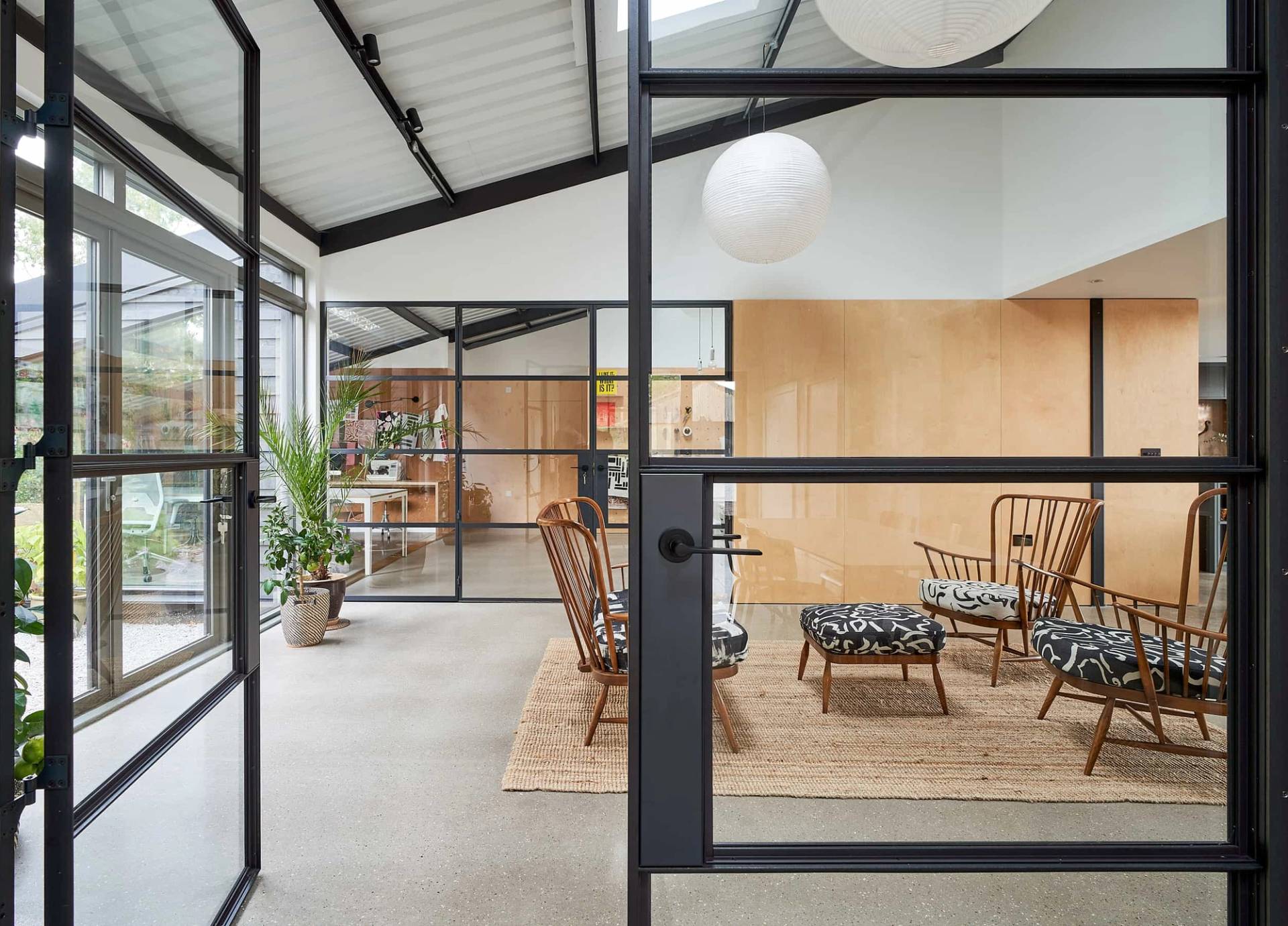 Crittall glass screen doorway looking into a light-filled lounge with woven armchairs and polished concrete floors, Love Locations IP30.