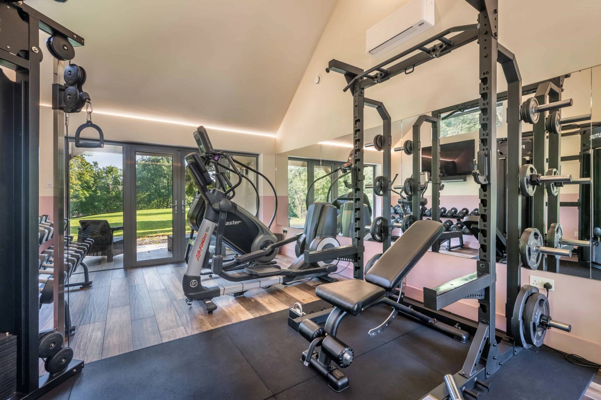 Fully equipped home gym outbuilding featuring workout racks, cardio equipment, and sliding glass doors opening to green gardens, ideal for fitness brand advertising, Love Locations.