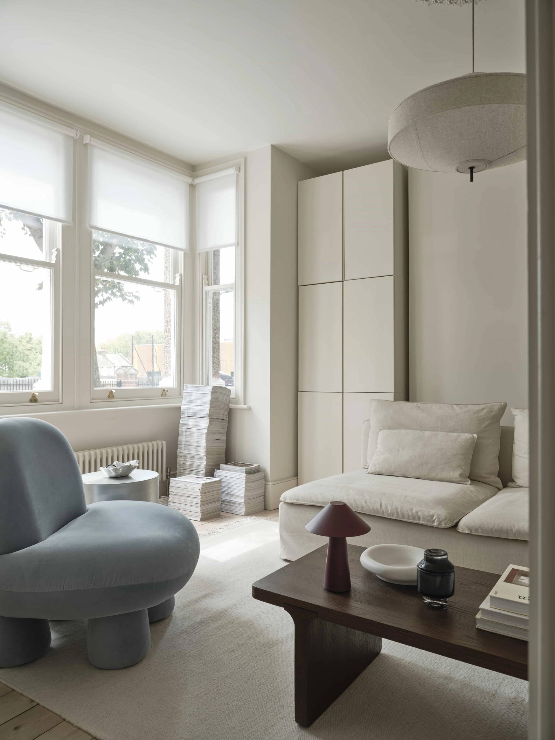 Minimalist lounge area with a light blue accent armchair, low cream sofa, and sheer window blinds in East London, Love Locations.