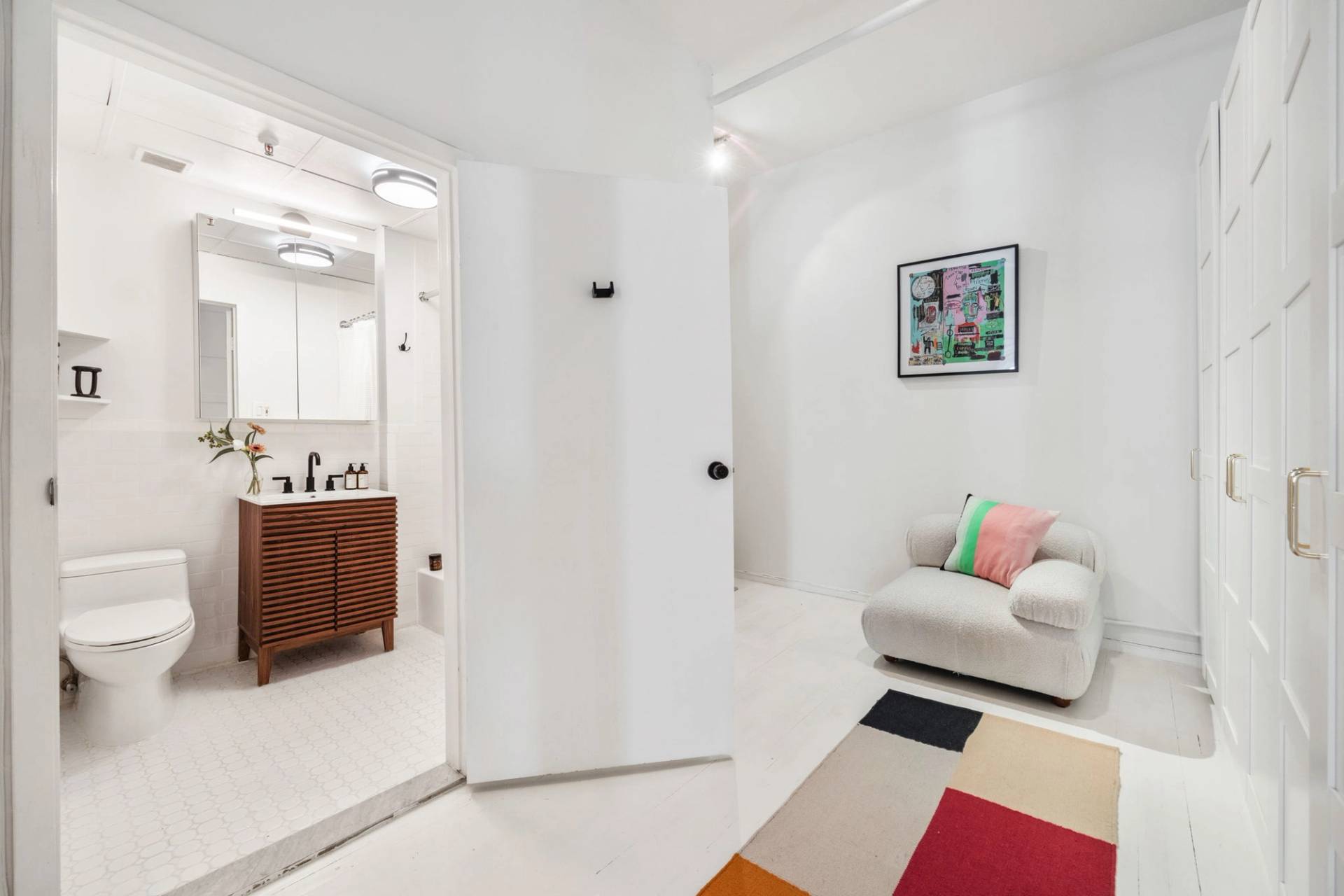 White entry hallway and seating alcove in a Tribeca artist loft film venue from Love Locations, featuring a cream armchair, colorful rug, and bathroom doorway.