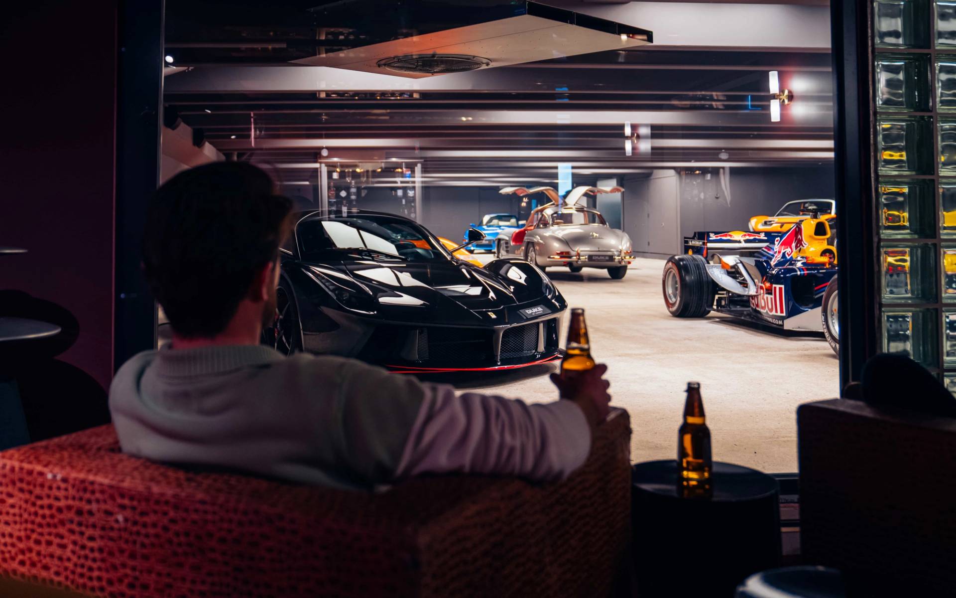 Intimate private bar seating nook with a guest holding a drink while viewing classic sports cars through a glass partition via Love Locations.
