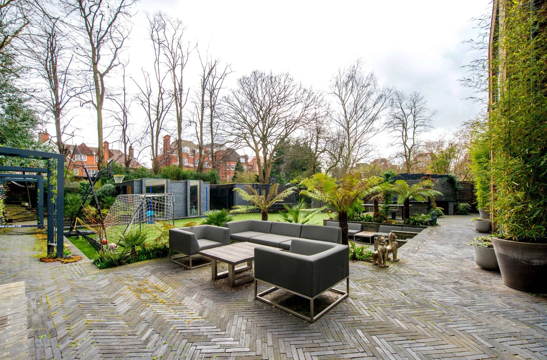 Private landscaped back garden showing outdoor lounge furniture, mature tropical planting, and a manicured lawn, Love Locations NW3.