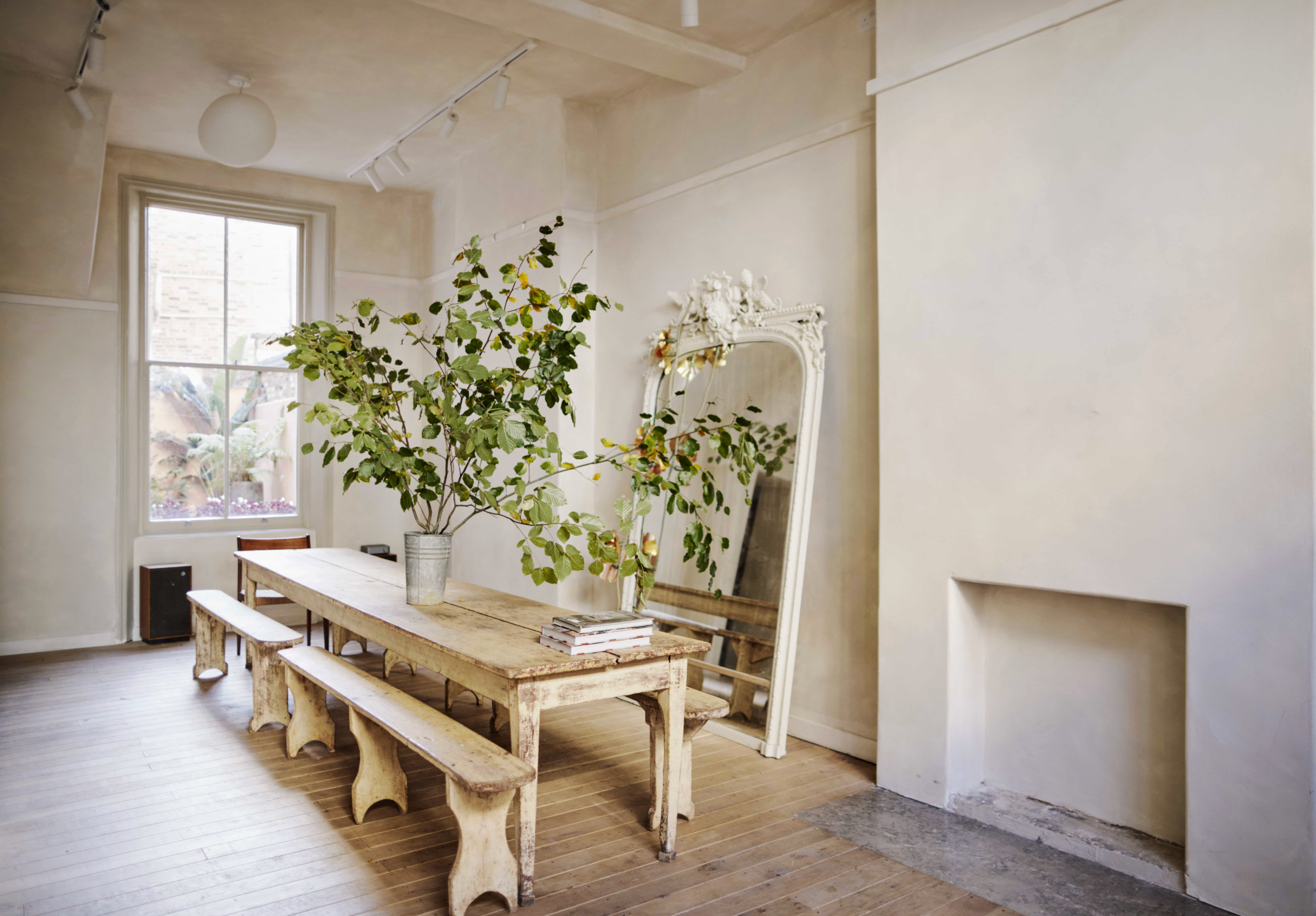 A light-filled dining room featuring a long rustic wooden table with matching benches, oversized leafy branches in metal buckets, an ornate leaning mirror, limewashed walls, and original timber floorboards.