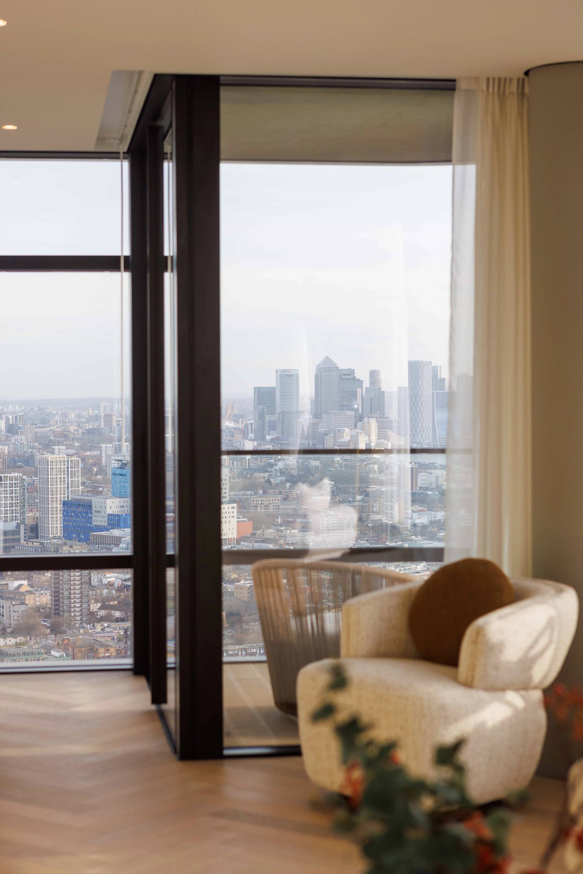 Cozy penthouse bedroom armchair reading corner set right against floor-to-ceiling glass framing London skyscraper views, ideal for quiet film scenes, Love Locations EC2A.