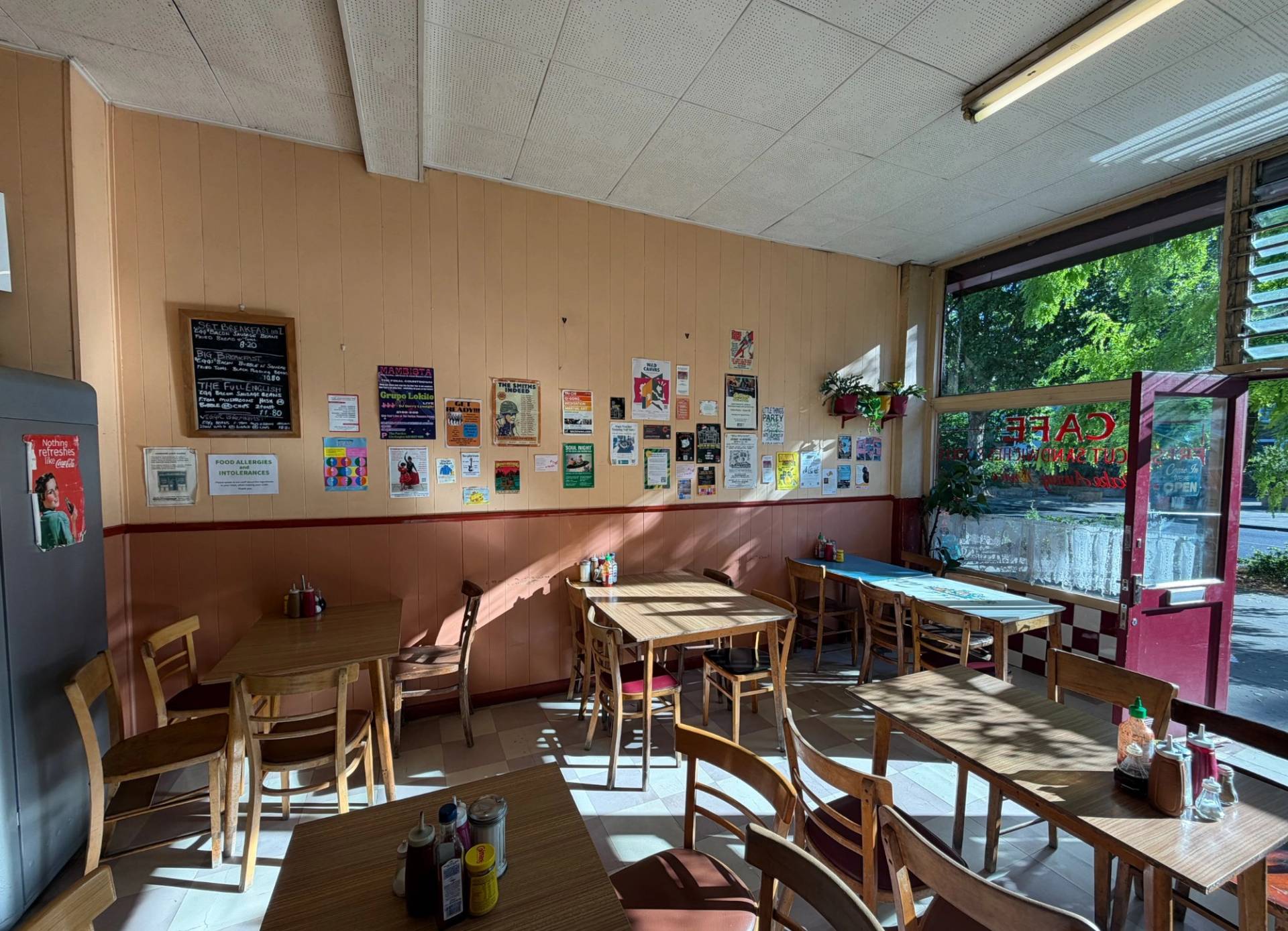 Classic cafe dining room interior with wooden tables, chairs, wall notices, and warm sunlight streaming through glass shopfront windows, Love Locations SE27.
