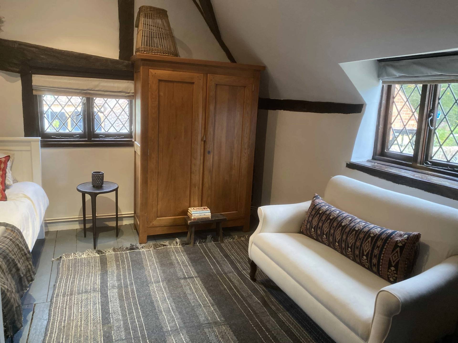 Cozy cottage bedroom seating nook featuring a white sofa and a large wooden wardrobe under historic sloping rafters, Love Locations.
