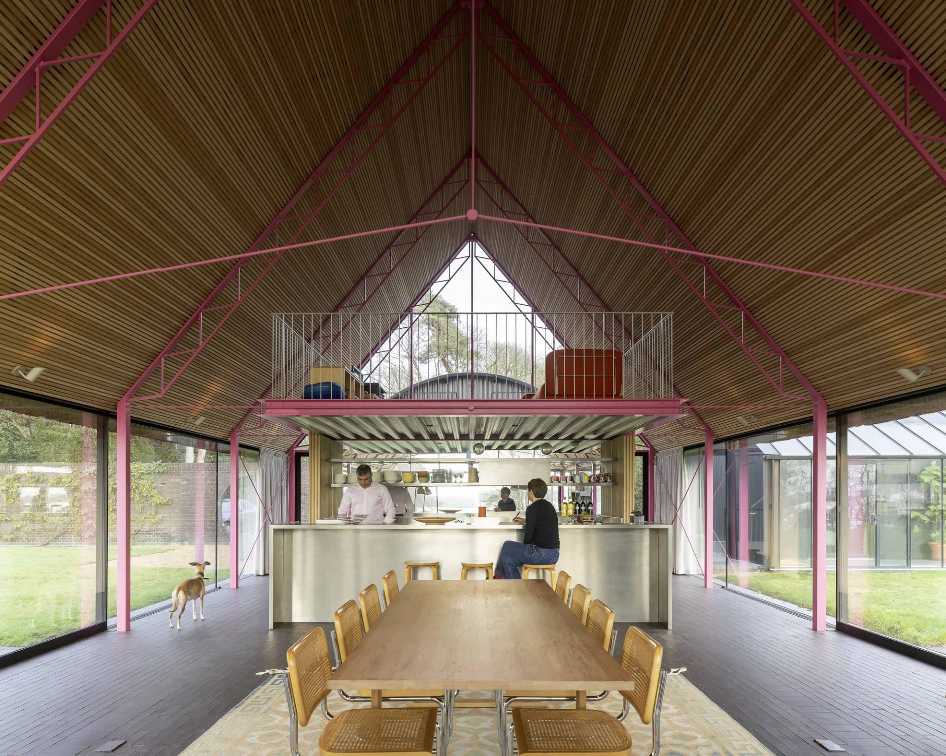 Double-height glass longhouse hall in IP17 with pink steel roof trusses, solid wood dining table, and open commercial kitchen, Love Locations Saxmundham.