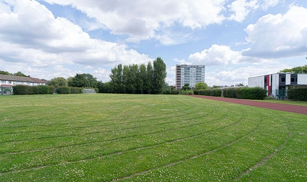 Love Locations outdoor athletics location South East London SE2: Large green school field featuring marked circular running lanes and a distant apartment block backdrop, perfect for athletic training ads.