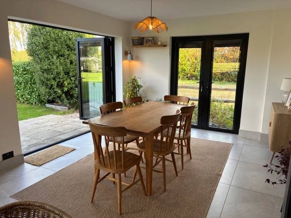 Bright dining room location house in Essex CM4 available through Love Locations, with a wooden dining set, warm brass pendant light, and black-framed bi-fold doors leading outside.