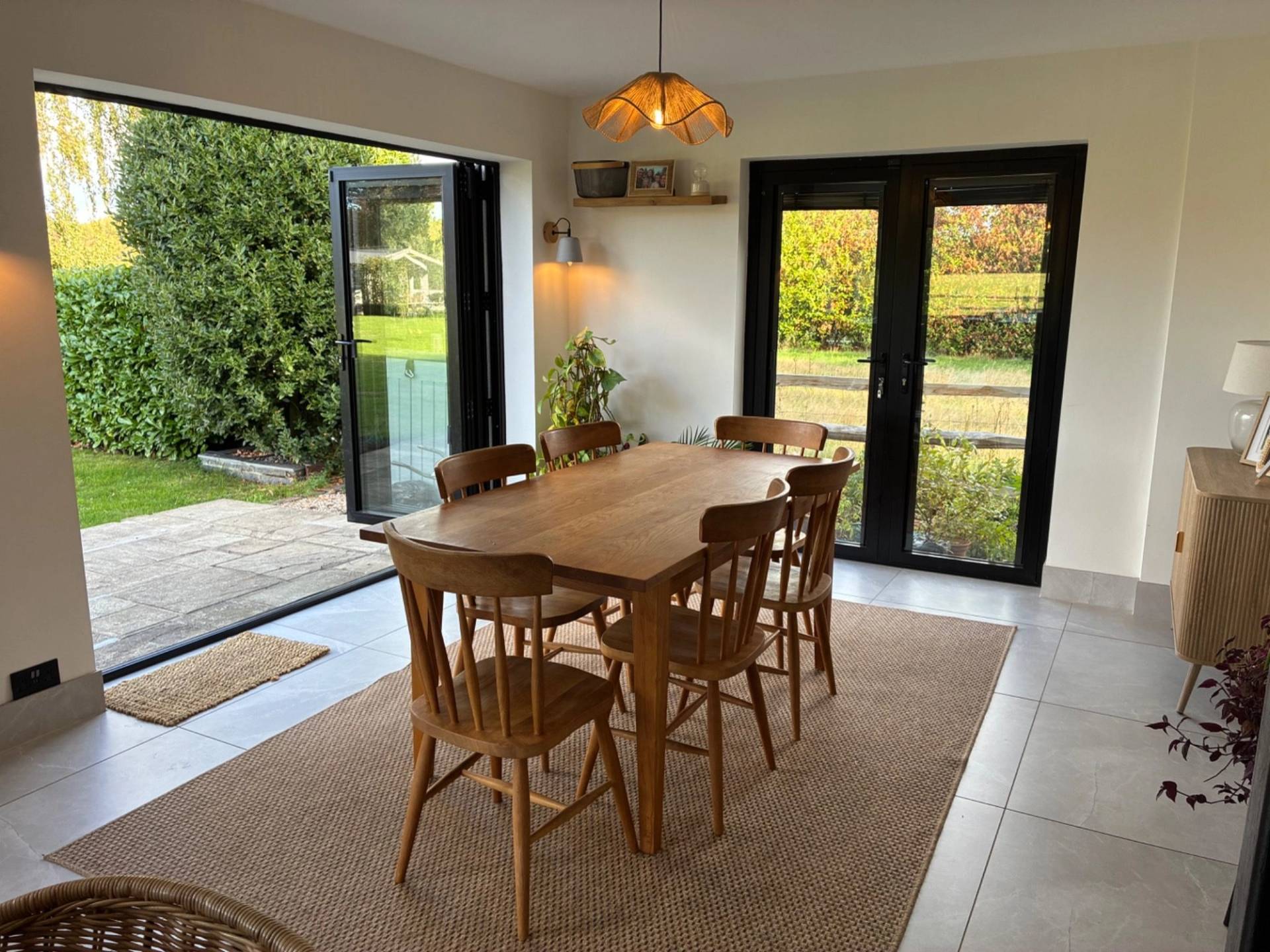 Bright dining room location house in Essex CM4 available through Love Locations, with a wooden dining set, warm brass pendant light, and black-framed bi-fold doors leading outside.
