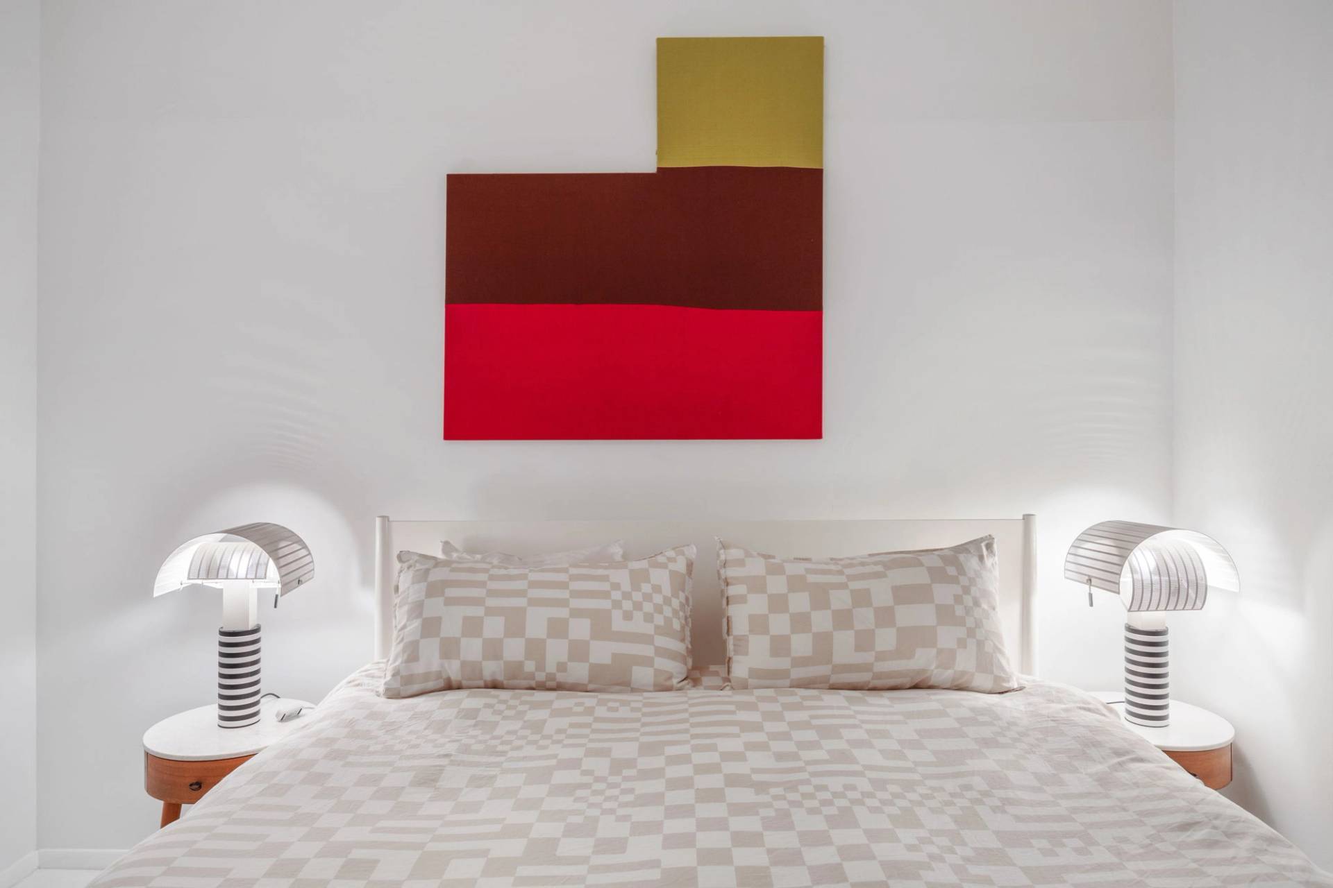 Close-up detail of a minimalist white bed with beige checkered bedding and color-blocked red wall art in a Tribeca loft shoot space from Love Locations.