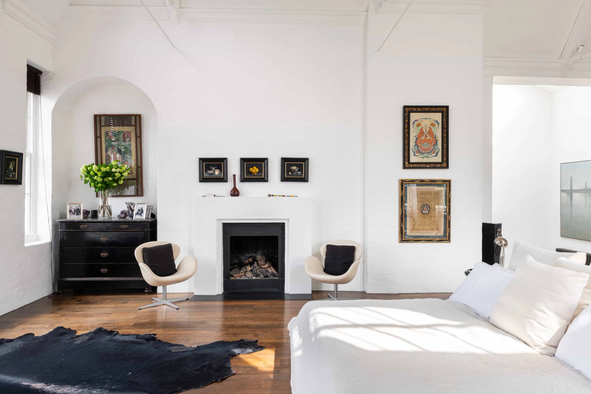 Master bedroom fireplace wall with a black hearth, twin mid-century chairs, and dark wood floors set under high ceilings, Love Locations SW11.