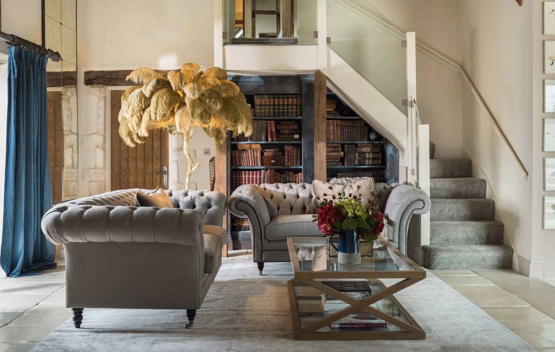 Double-height reception lounge shoot venue in Cheltenham GL51 via Love Locations, featuring plush grey tufted Chesterfield sofas, glass balustrade mezzanine, and a dramatic pampa grass display.