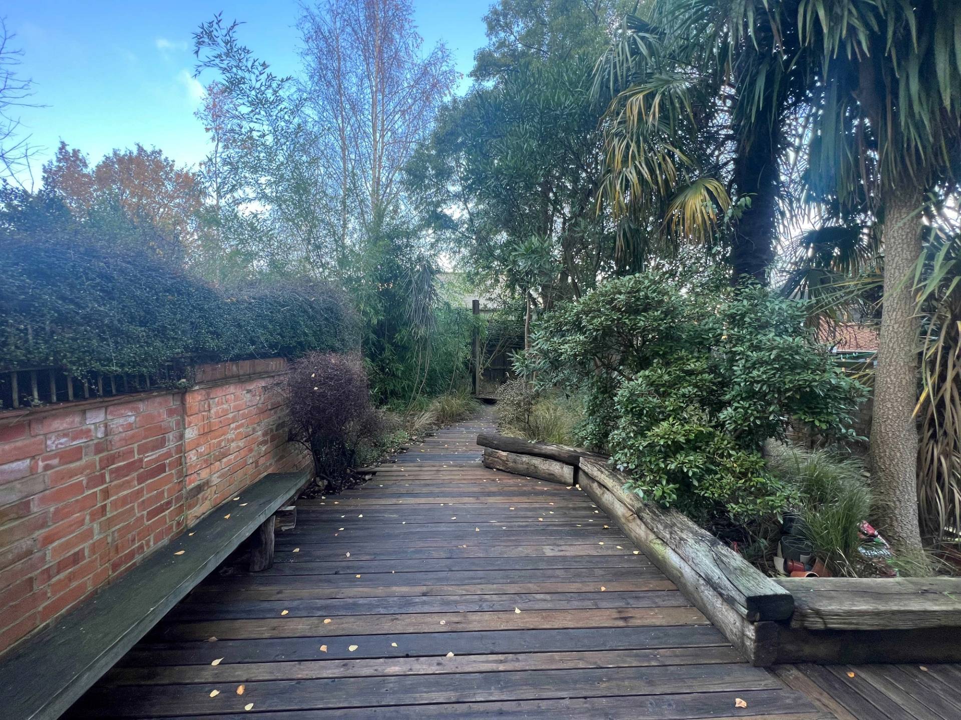 Secluded rear garden walkway with wooden decking planks, raised timber borders, and lush mature greenery, Love Locations Denmark Hill.