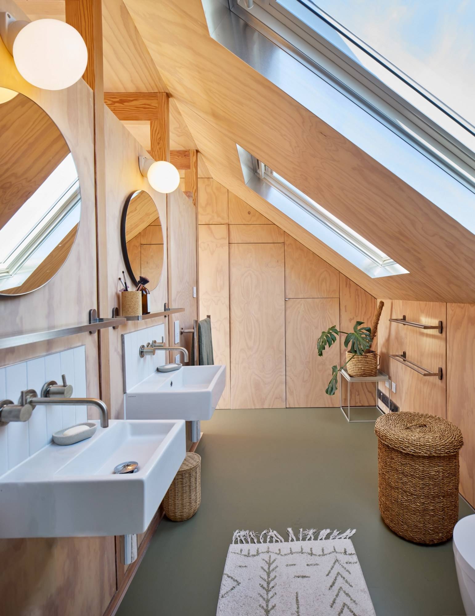 Loft converter bathroom featuring dual white pedestal sinks, round mirrors, skylights, and olive green rubber flooring, Love Locations E17.