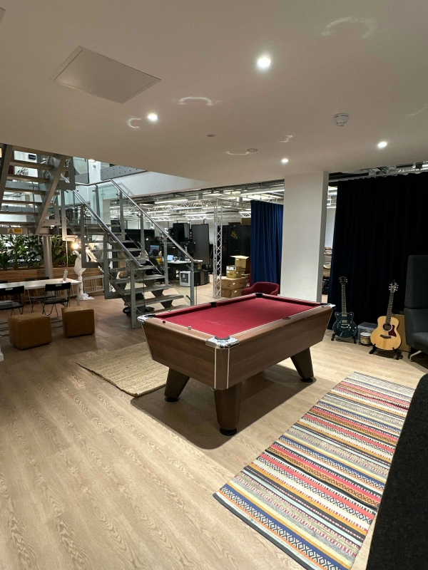 Bright open office space with industrial-style exposed ceiling, pale wooden floorboards, and a social breakout area featuring a red baize-covered pool table, colourful striped rug, and a guitar.