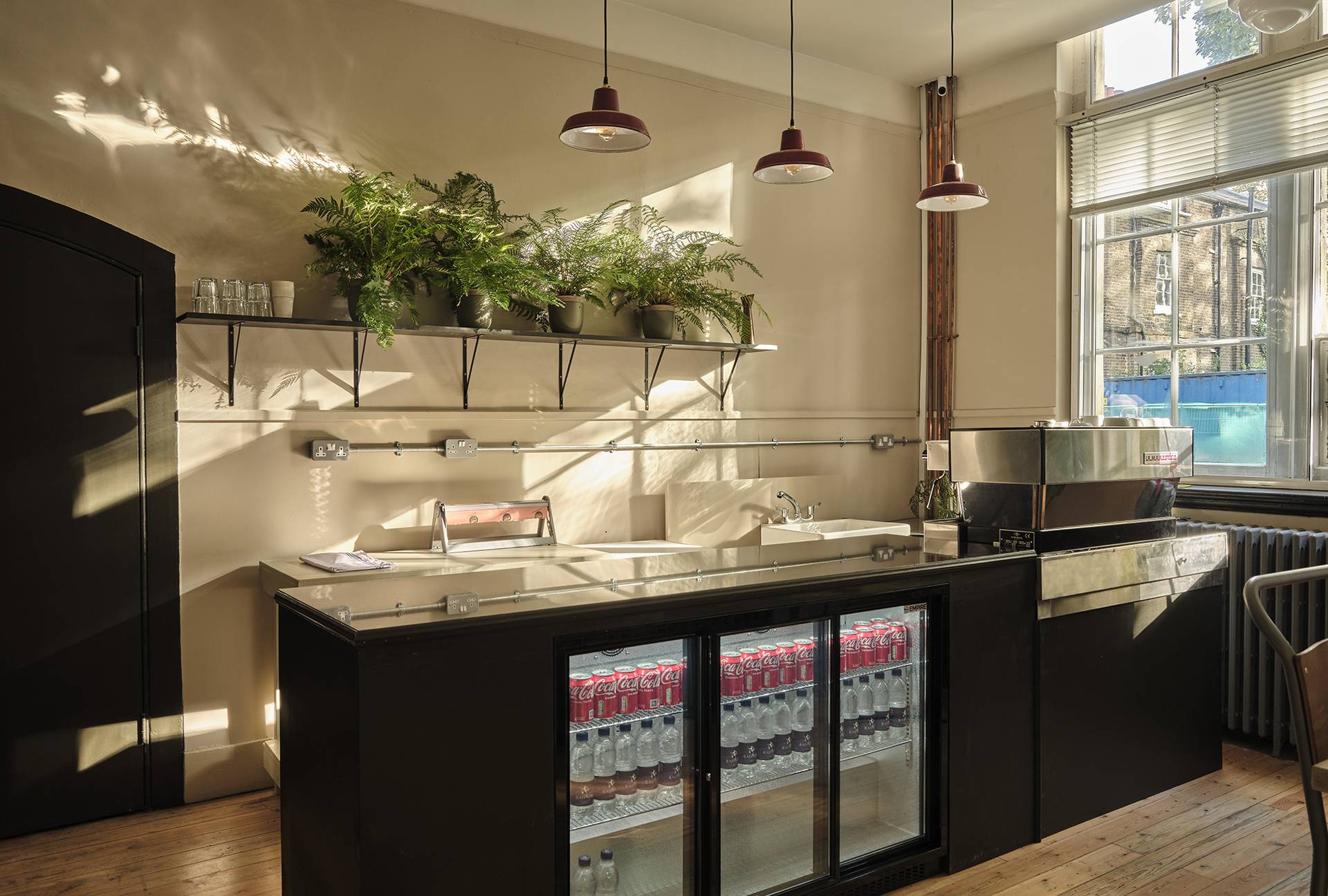 Modern bar counter area featuring under-counter glass front fridges, open shelving, and potted plants, perfect for hospitality filming, Love Locations E2.