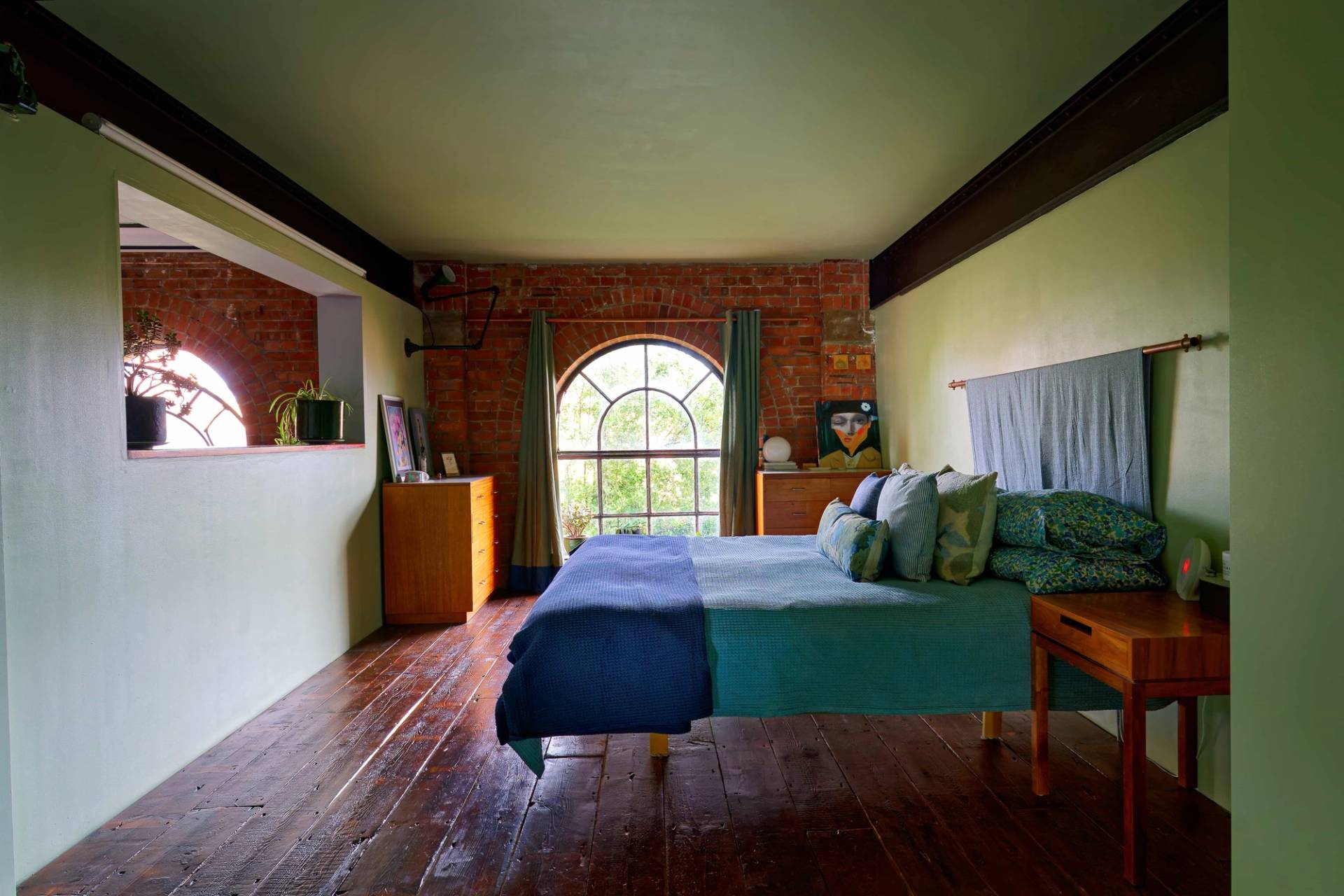 Warehouse bedroom suite featuring exposed red brick walls, arched window, dark timber floors, and blue bed throw, Love Locations E14.