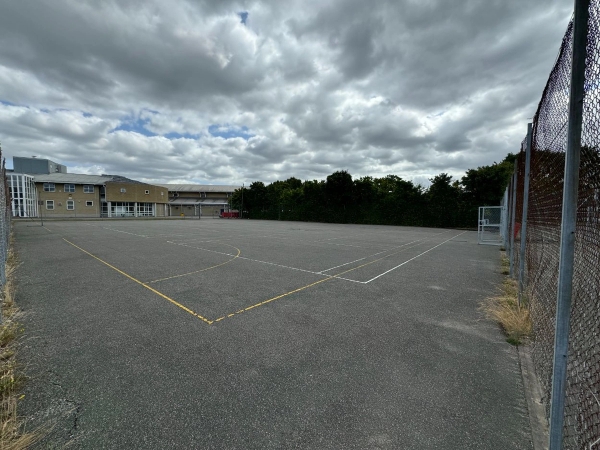 Love Locations open school playground location EN4: Wide-open outdoor tarmac games court with painted boundary lines under a dramatic cloud sky, perfect for urban youth dramas.