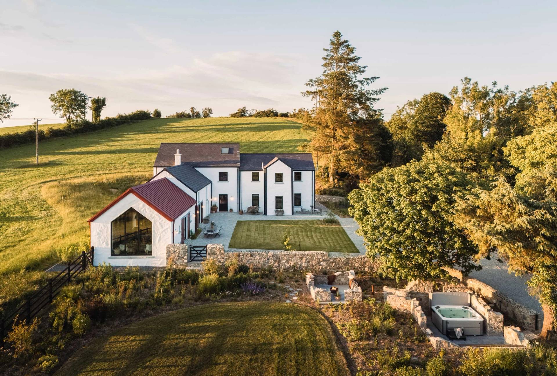 High aerial view of a restored luxury farmhouse estate film location in Castleblayney County Monaghan Ireland from Love Locations, showcasing white stone buildings, red roof accents, a private lawn, sunken fire pit, hot tub, and surrounding green Irish fields.