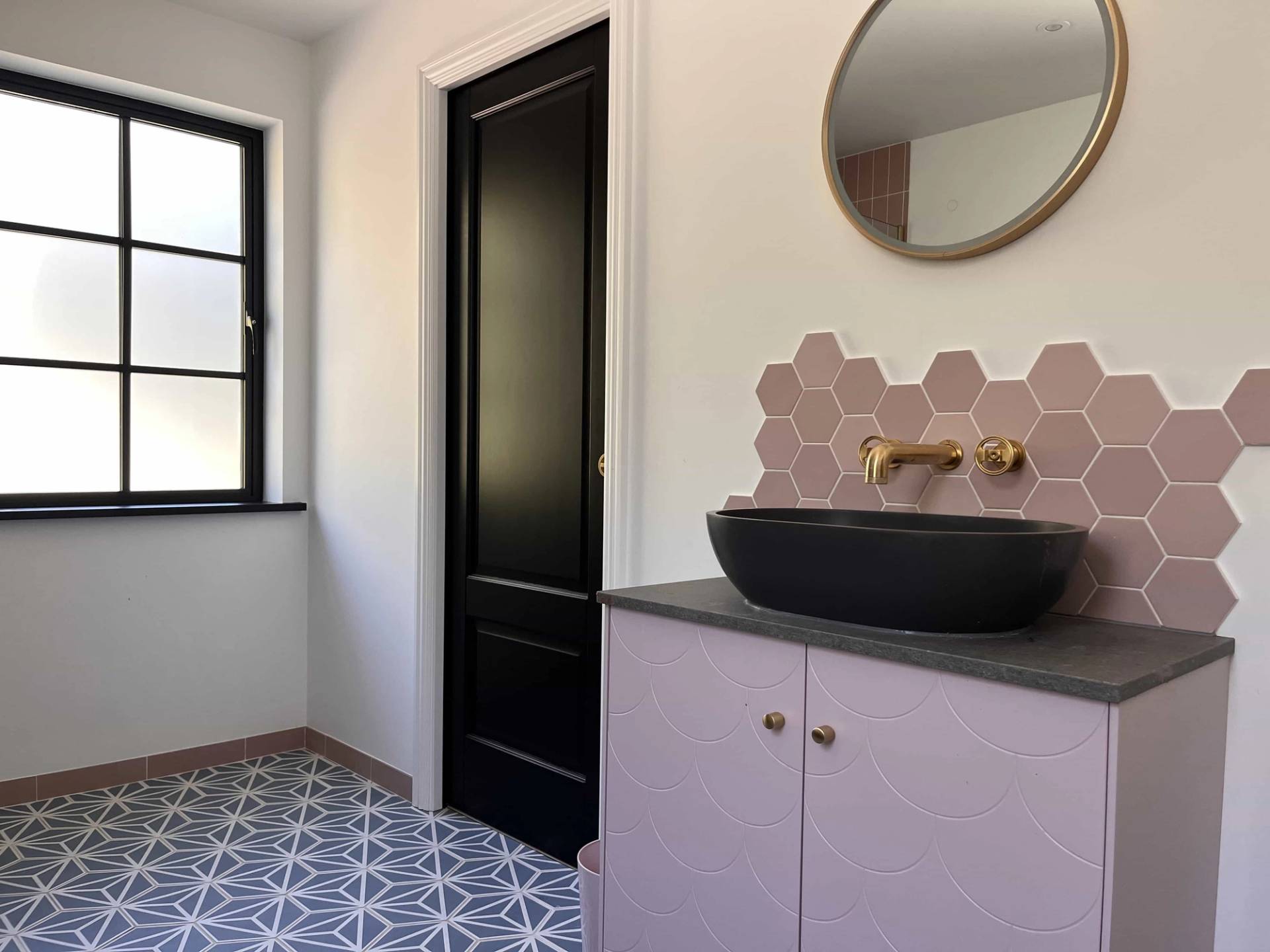Powder room vanity featuring pink hexagon tiles, round wall mirror, black vessel sink, and patterned tiles, Love Locations GU3.
