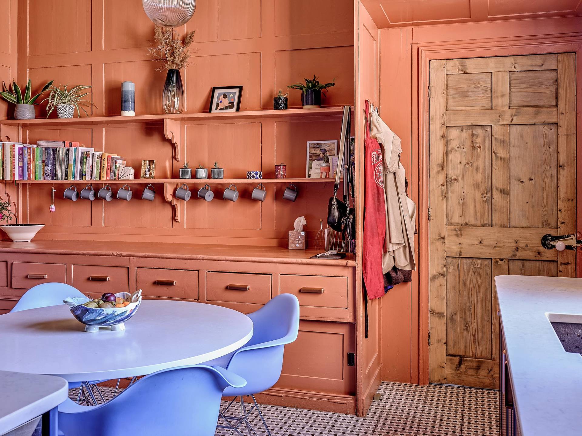 Terracotta orange kitchen dining corner featuring built-in dresser shelving, blue retro chairs, and a rustic wooden door, Love Locations.