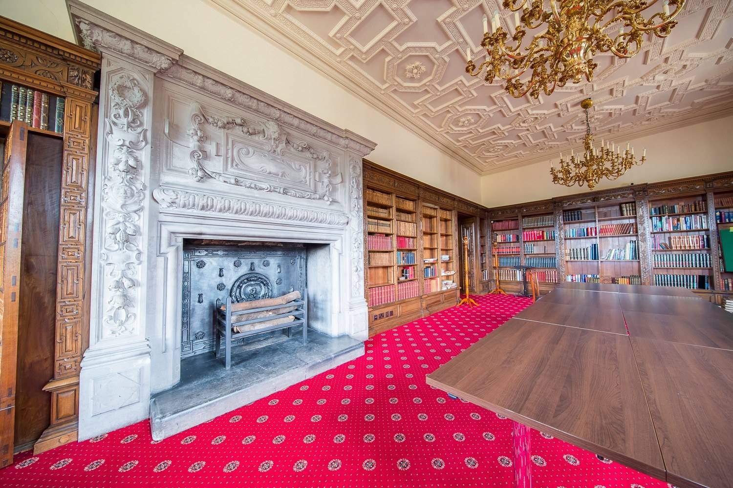 Stately library location house in Hertfordshire AL9 through Love Locations, featuring wall-to-wall fitted bookcases, grand carved mantlepiece, red patterned carpet, and ornate plasterwork ceiling.
