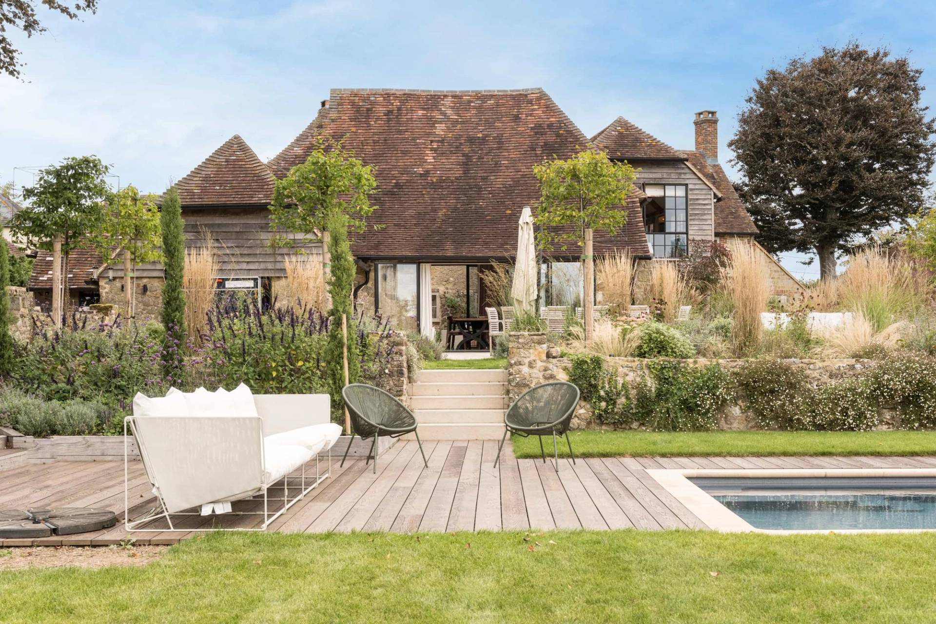 Converted stone barn elevation with timber decking, swimming pool, and outdoor sun lounger terrace, Love Locations GU28. Paved