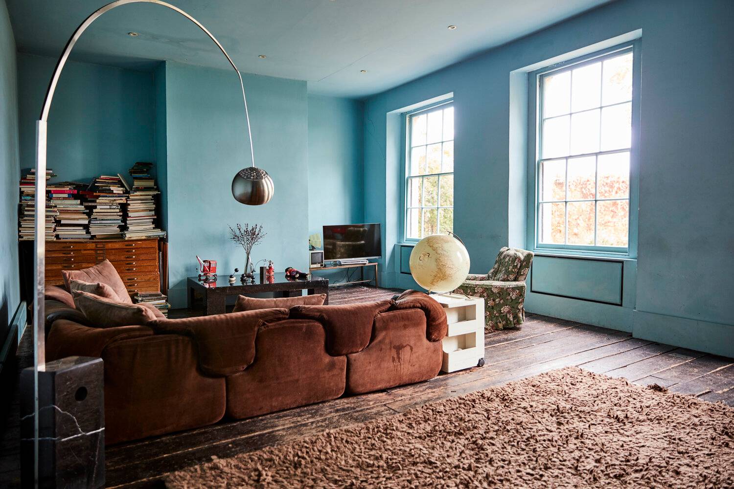 Sky blue reception room featuring a curved brown velvet sectional sofa, arc floor lamp, and tall sash windows, ideal for lifestyle campaigns, Love Locations SE8.