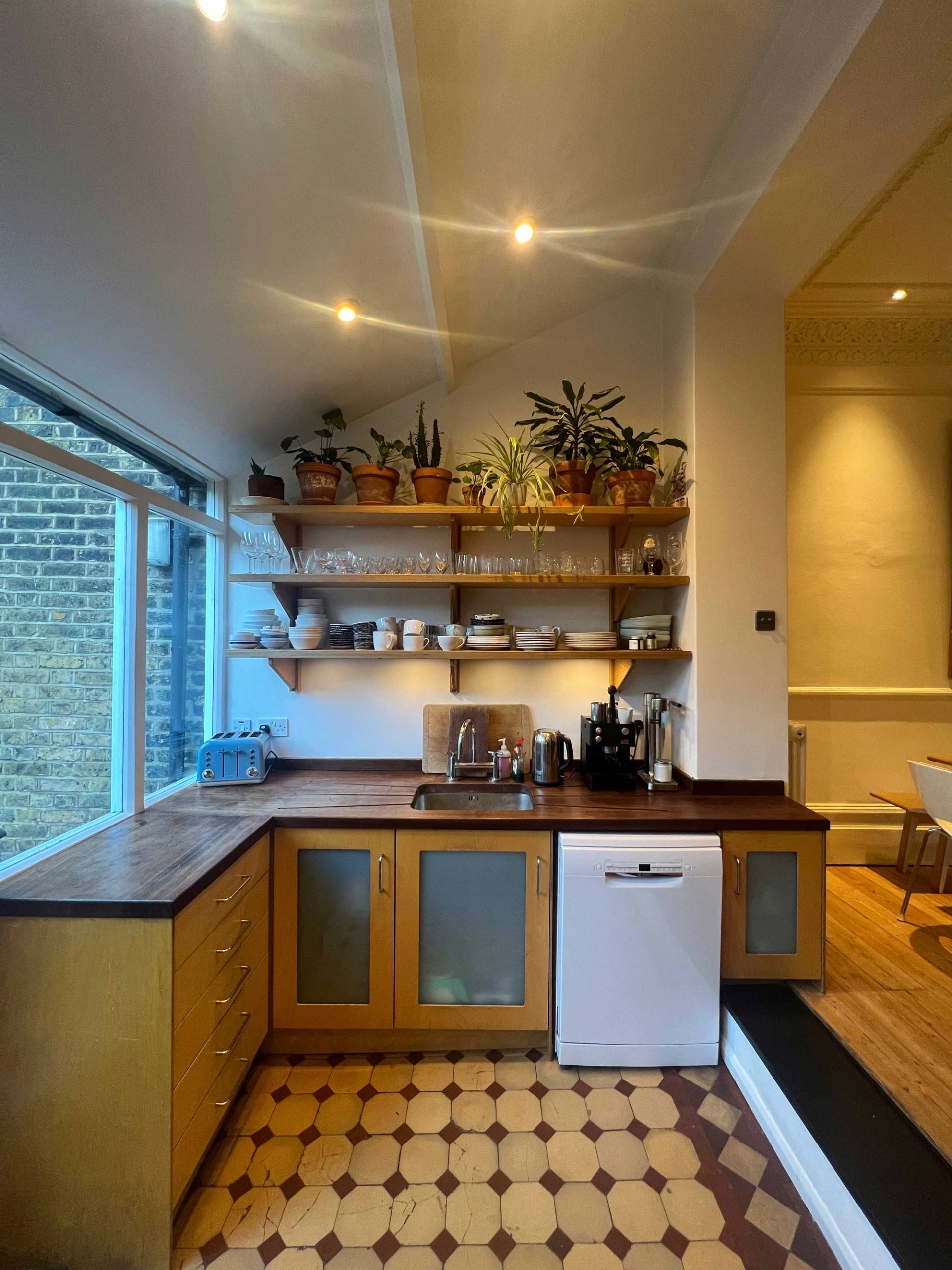 Galley kitchen view showcasing open timber wall shelves filled with potted plants and ceramics set above warm wooden counters, Love Locations SE5.
