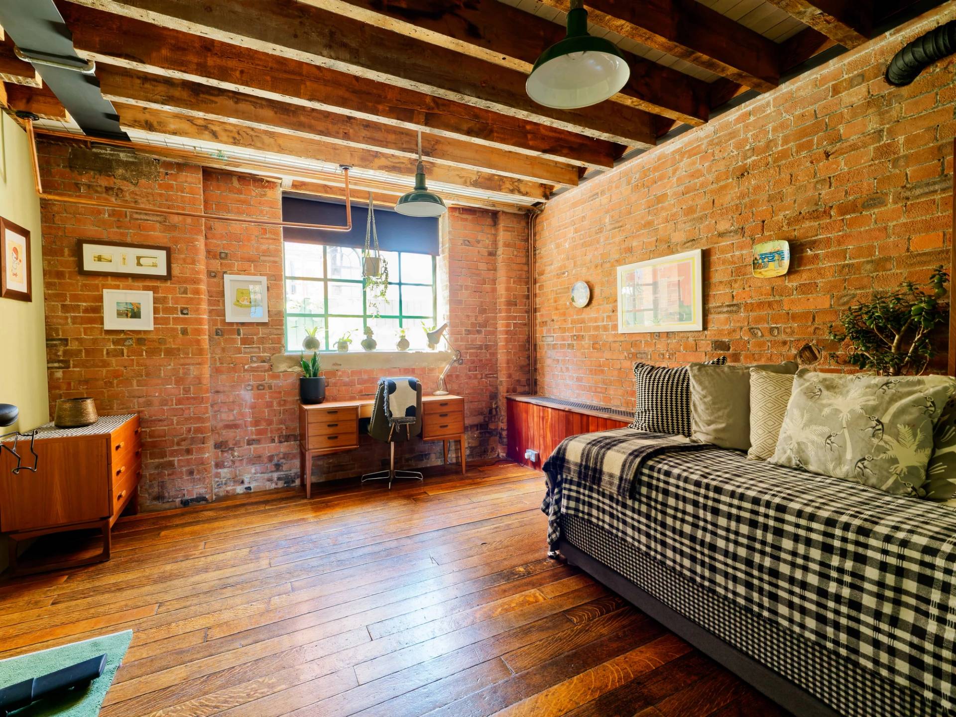 Industrial bedroom featuring exposed brickwork, timber ceiling beams, dark wood floors, and plaid sofa, Love Locations E14.