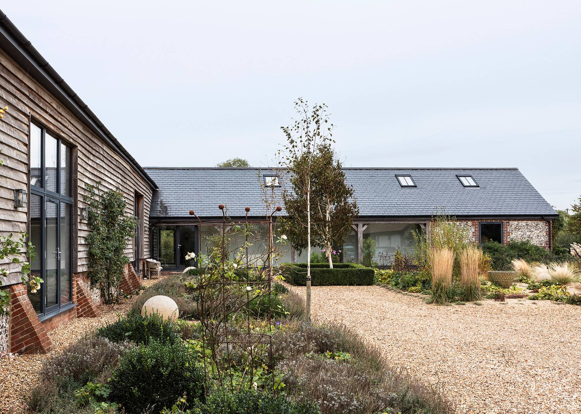 Gravel entrance courtyard lined with stone paving, native shrubs, and weatherboard barn architecture, Love Locations SN8.