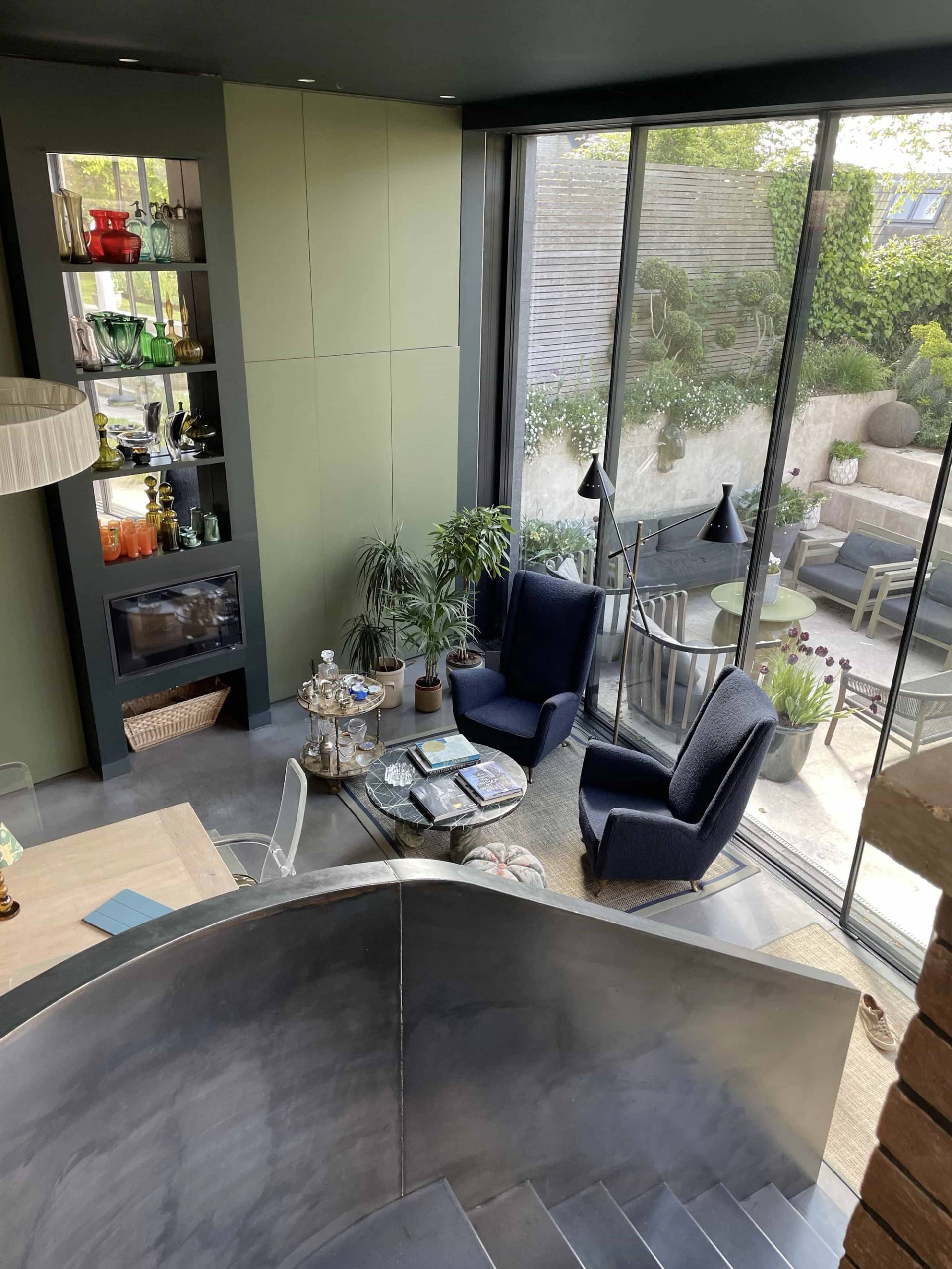 High-angle bird's-eye view from a modern steel staircase into an open-plan lounge area with plush armchairs and glass courtyard doors, Love Locations.