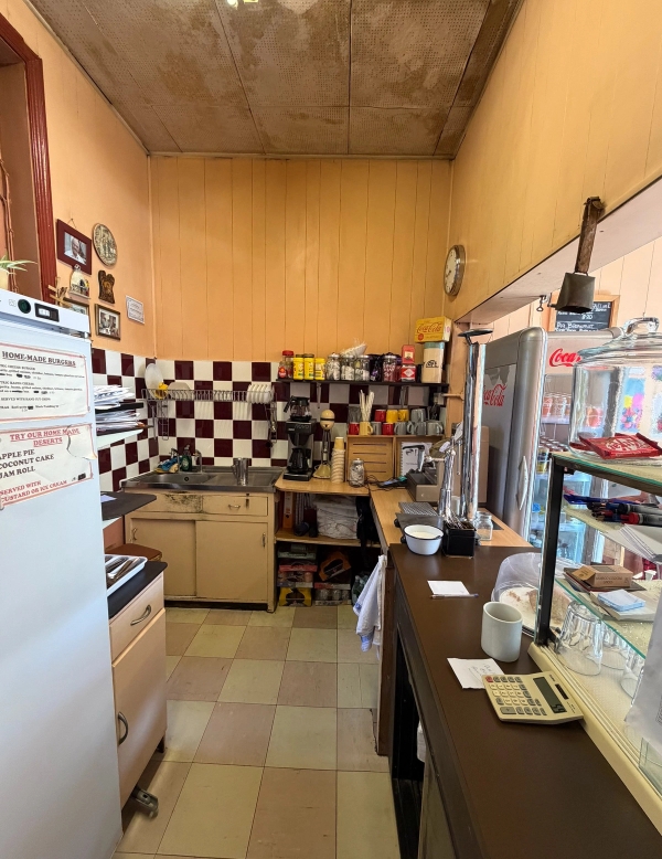 Behind-the-counter perspective of a classic London diner cafe showing food prep areas, shelving, and checkered tiles, Love Locations SE27.