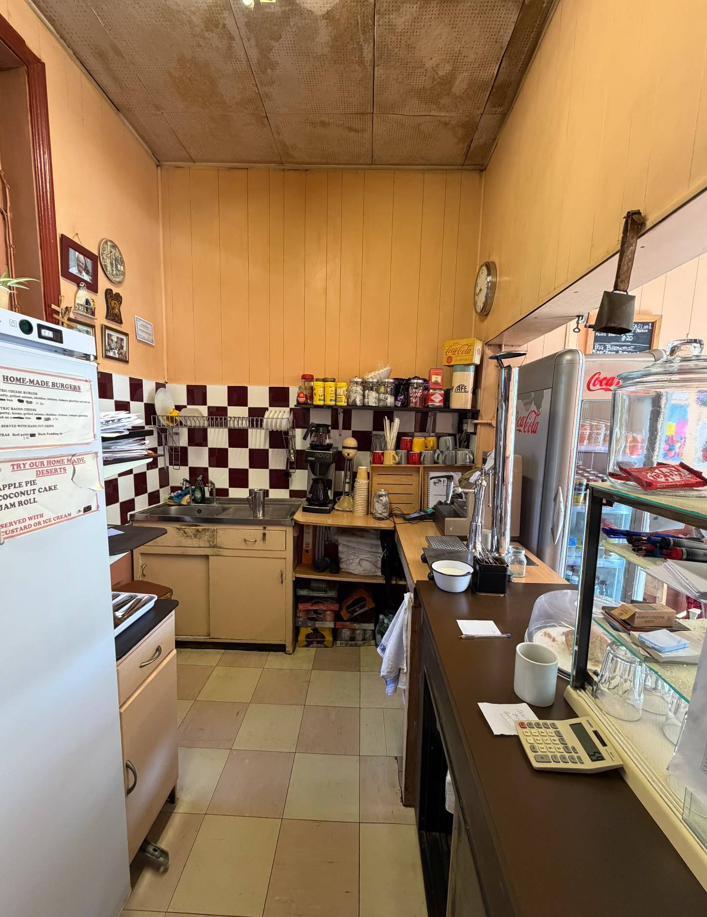 Behind-the-counter perspective of a classic London diner cafe showing food prep areas, shelving, and checkered tiles, Love Locations SE27.