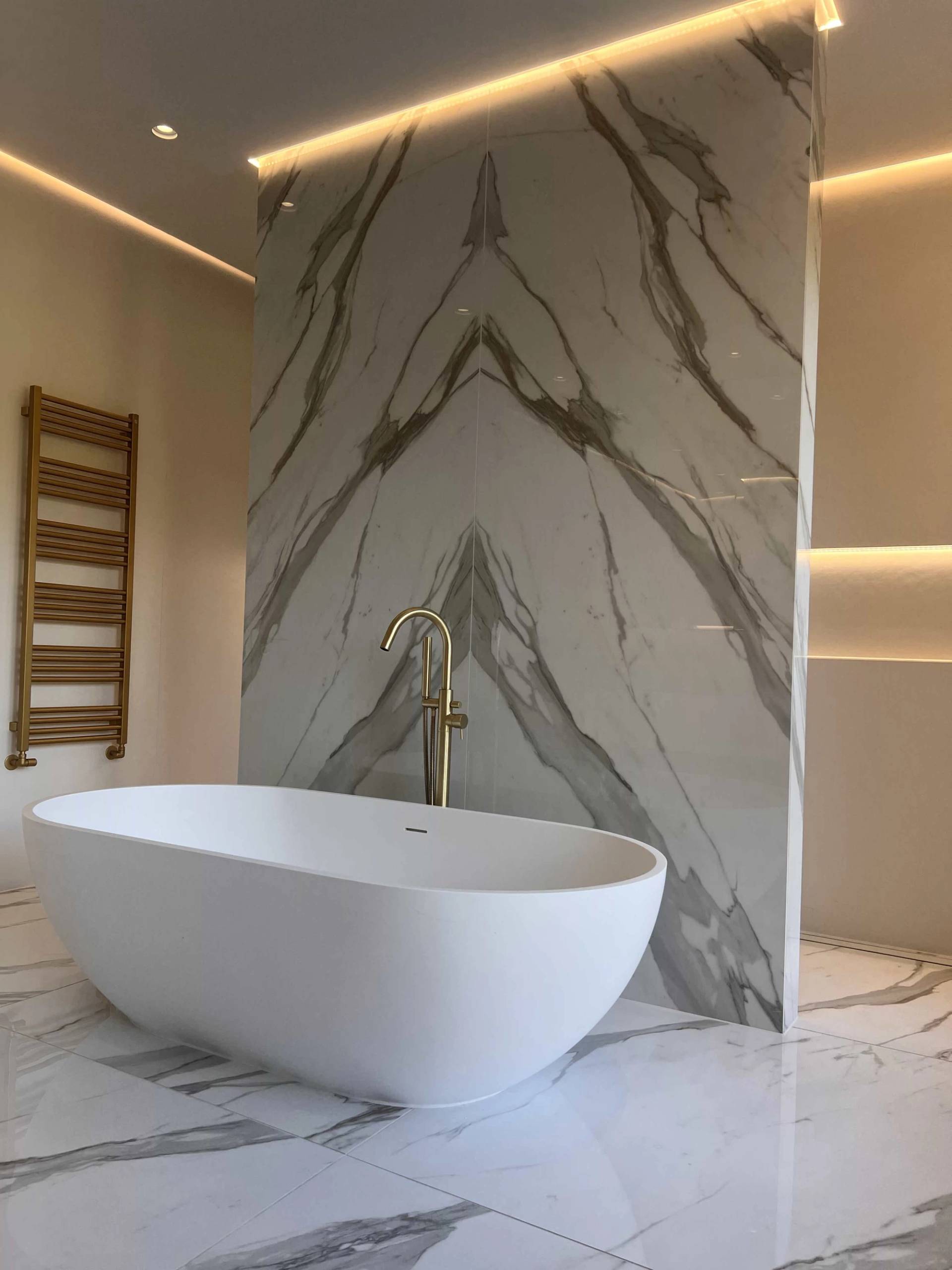 Close-up of a modern freestanding bathtub with gold floor-mounted tapware and book-matched marble tile accents, ideal for wellness product filming, Love Locations.