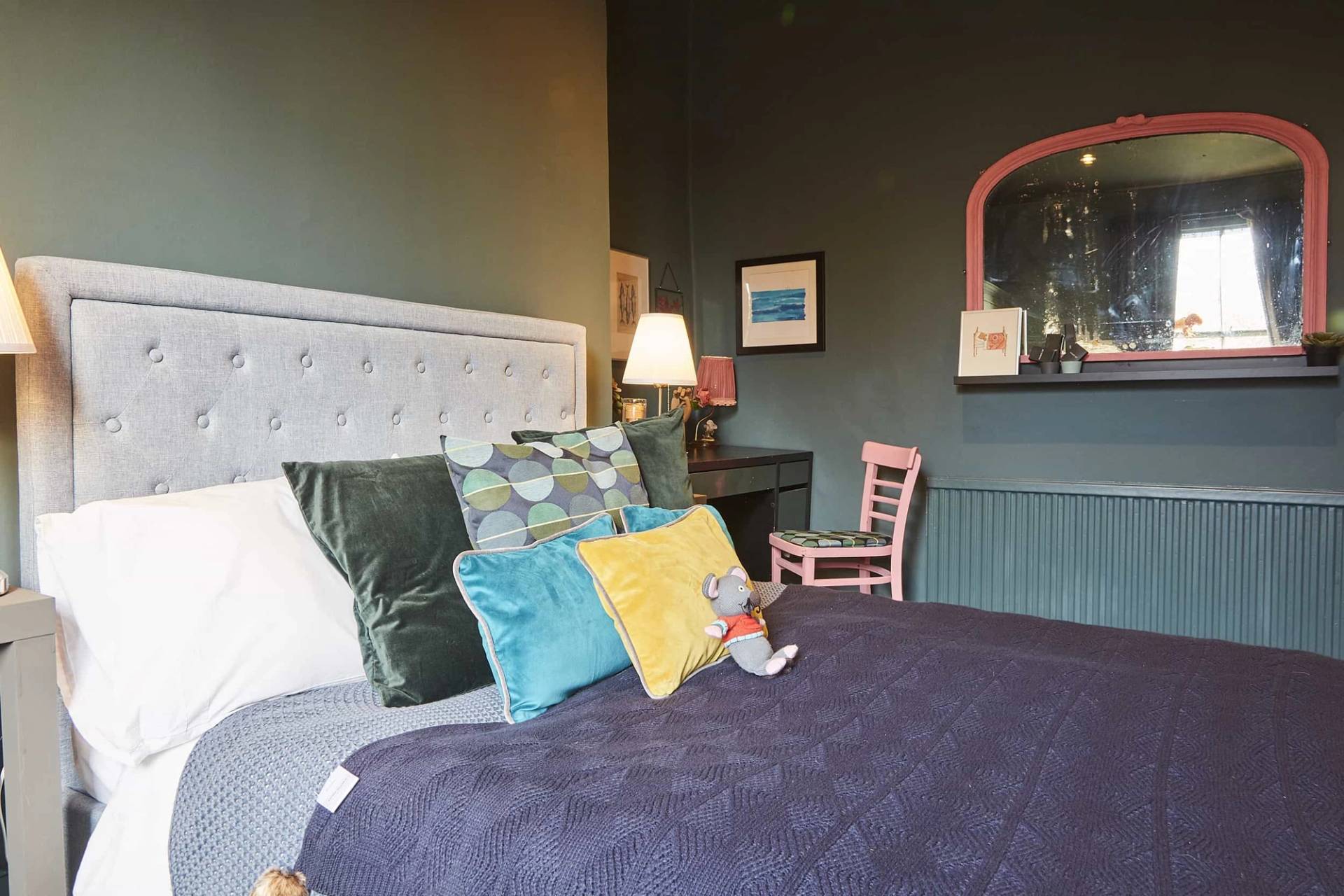 Dark teal bedroom setup featuring a white upholstered bed, colorful throw pillows, and warm bedside lamps, Love Locations N5.