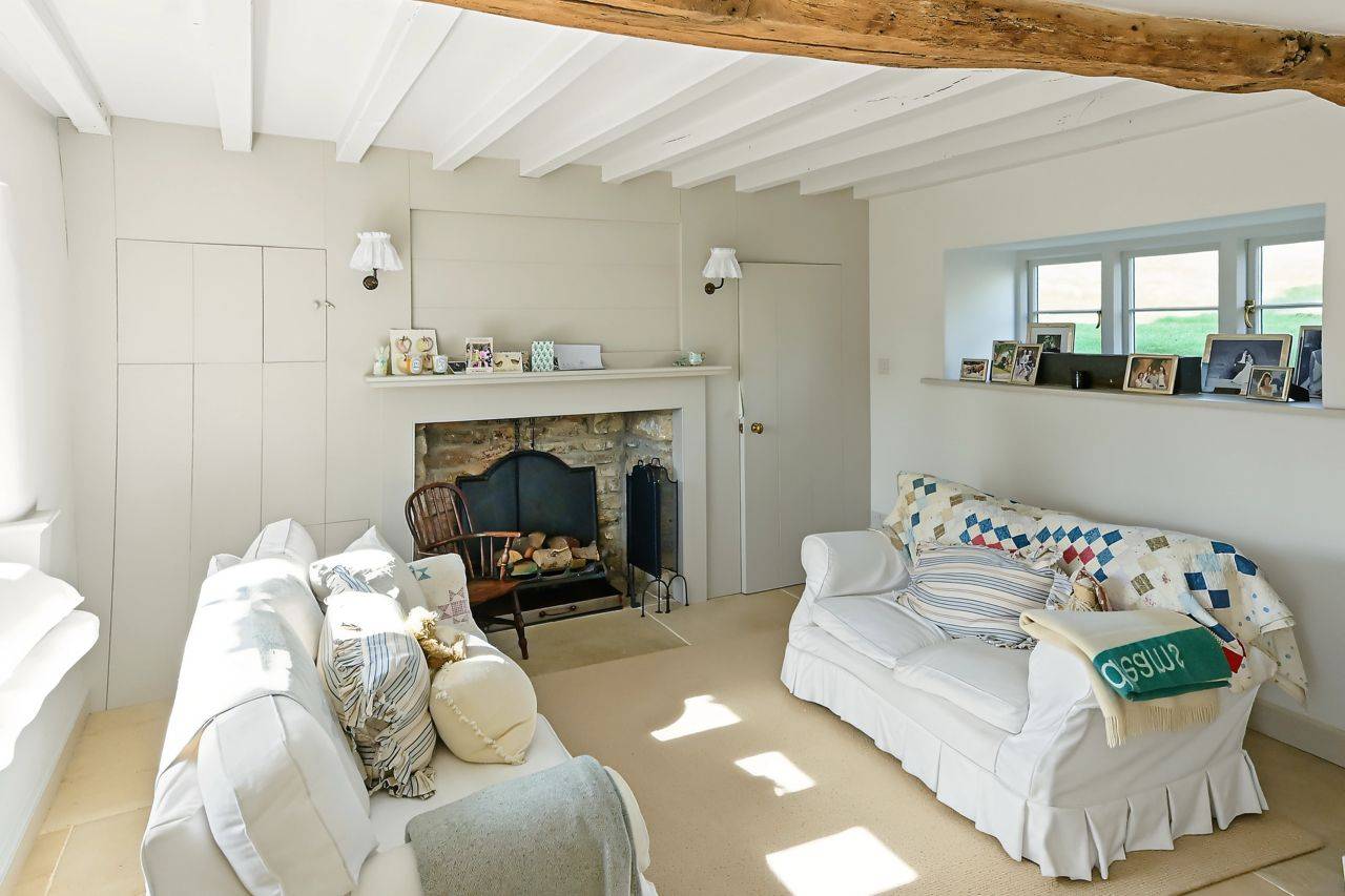 Cozy cottage sitting room shoot location in Gloucestershire GL7 via Love Locations, showcasing exposed wooden roof beams, a traditional stone fireplace, and soft cream furnishings.