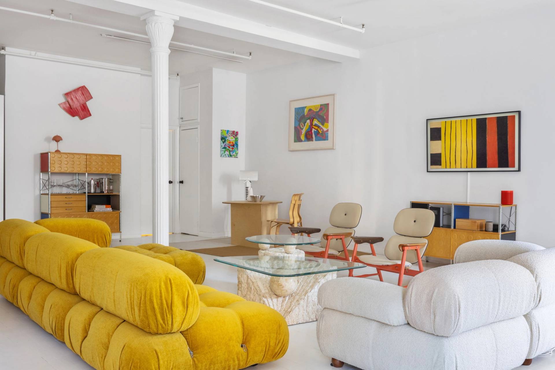 Bright loft sitting area with white cast-iron pillars, colorful artwork, glass coffee table, and mustard velvet sofa in Tribeca NYC via Love Locations.