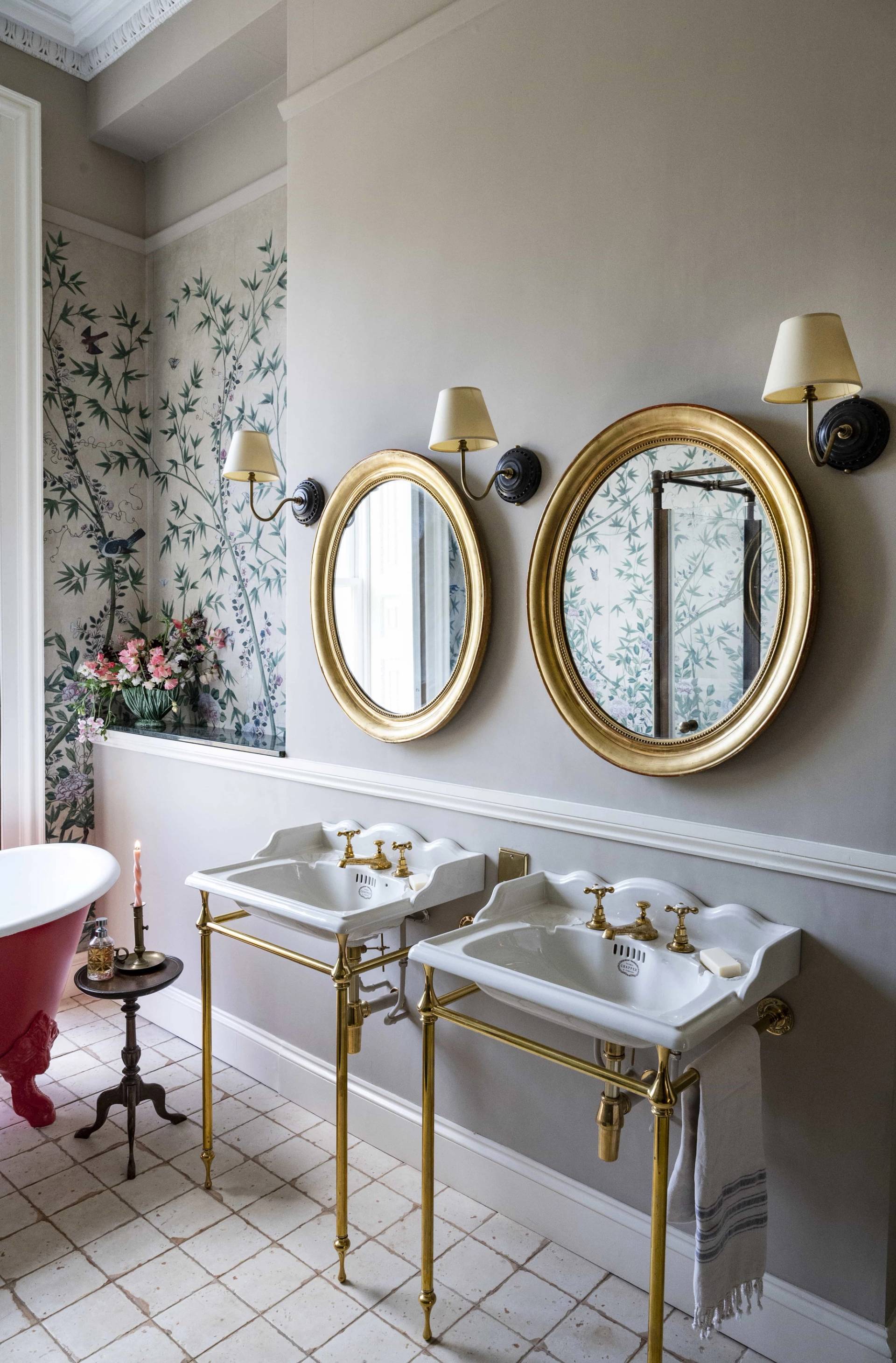 Dual oval brass mirrors and twin marble vanity sinks against subtle floral wallpaper in a manor house bathroom, Love Locations.