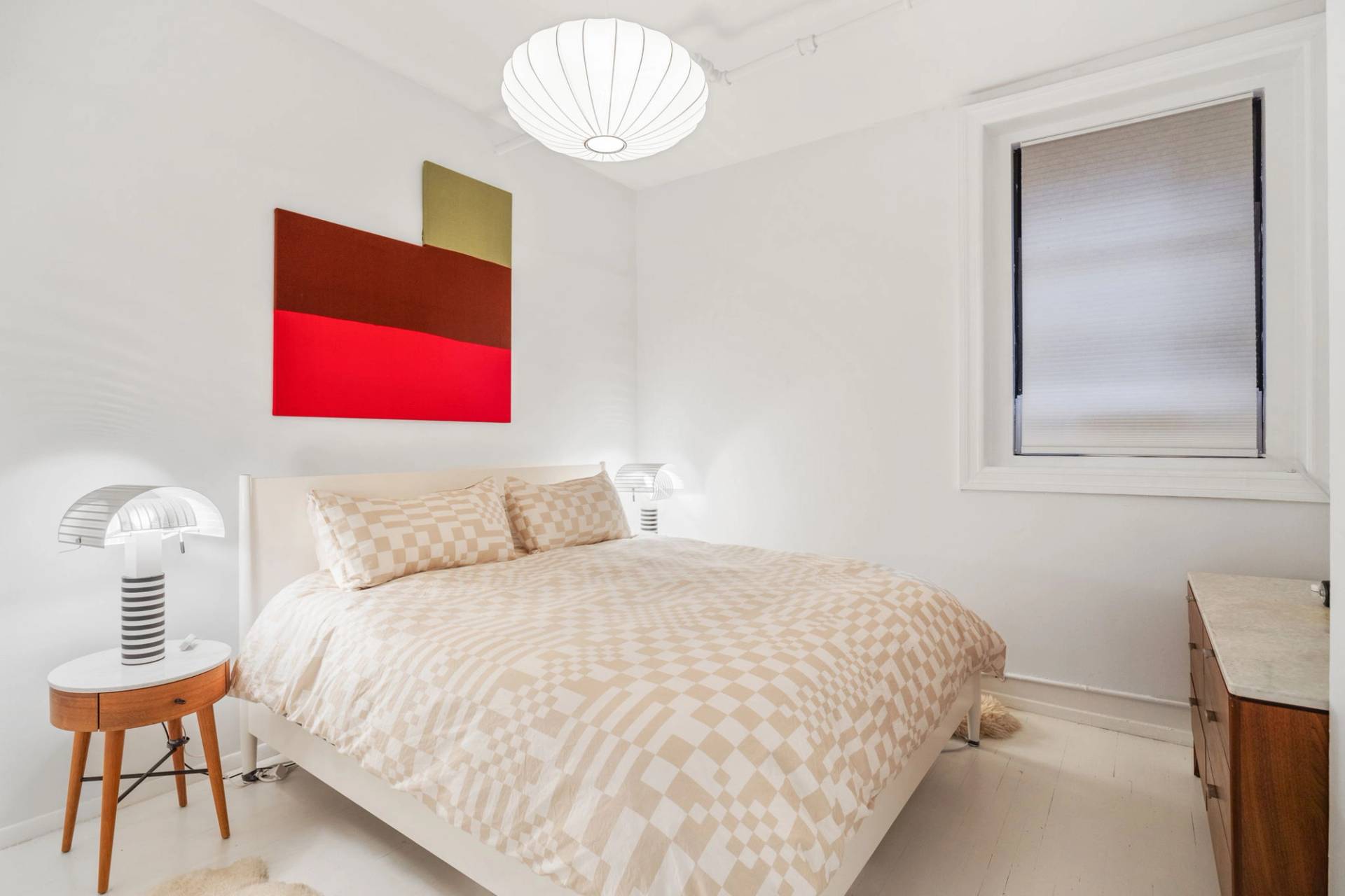 Minimalist master bedroom film location with a white bed frame, beige checkered duvet, red wall art, and paper pendant light in Tribeca NY 10007 via Love Locations.