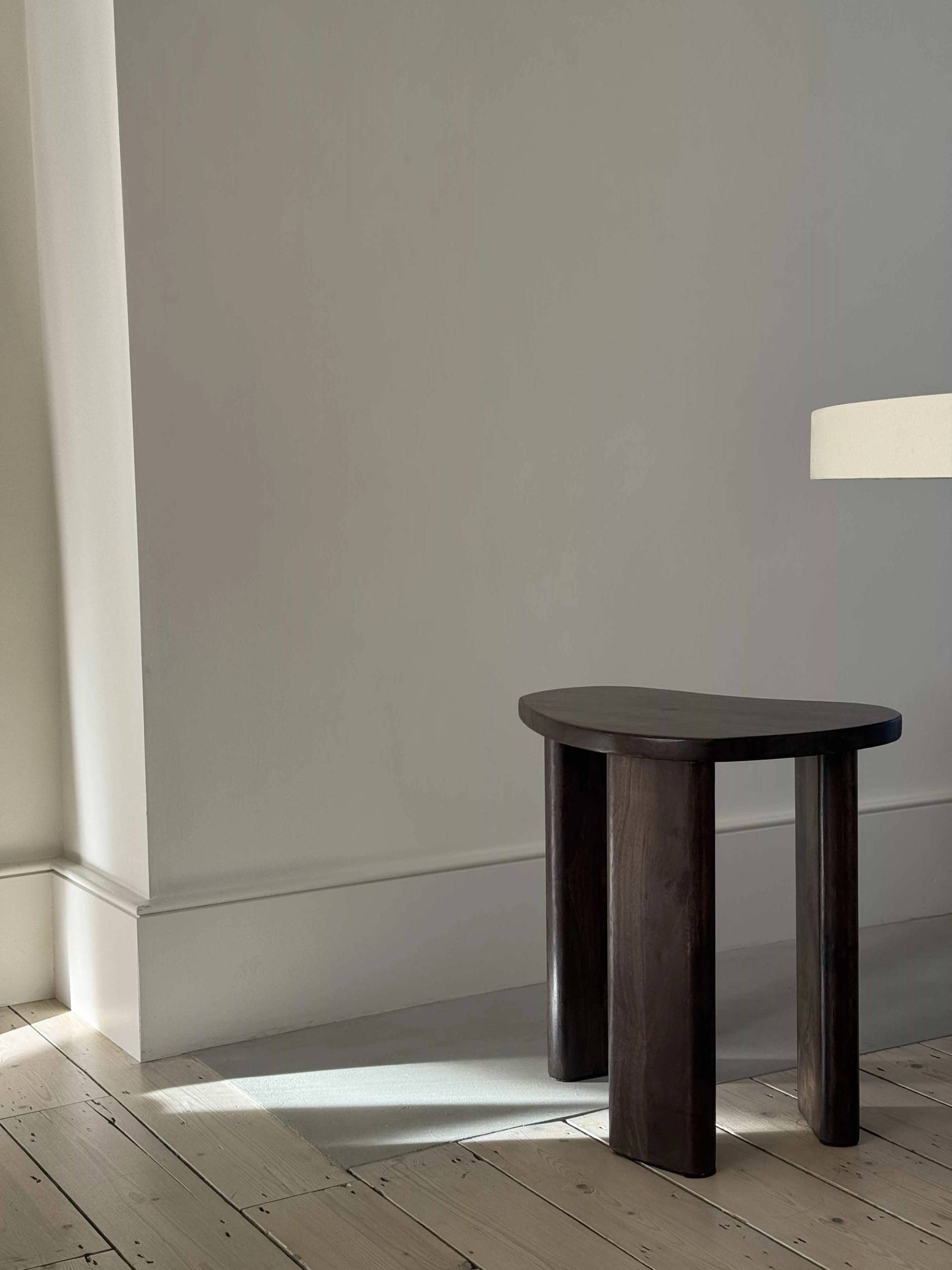 Minimalist room corner featuring a dark wood tripod side table cast in soft natural window sunlight, Love Locations E4.